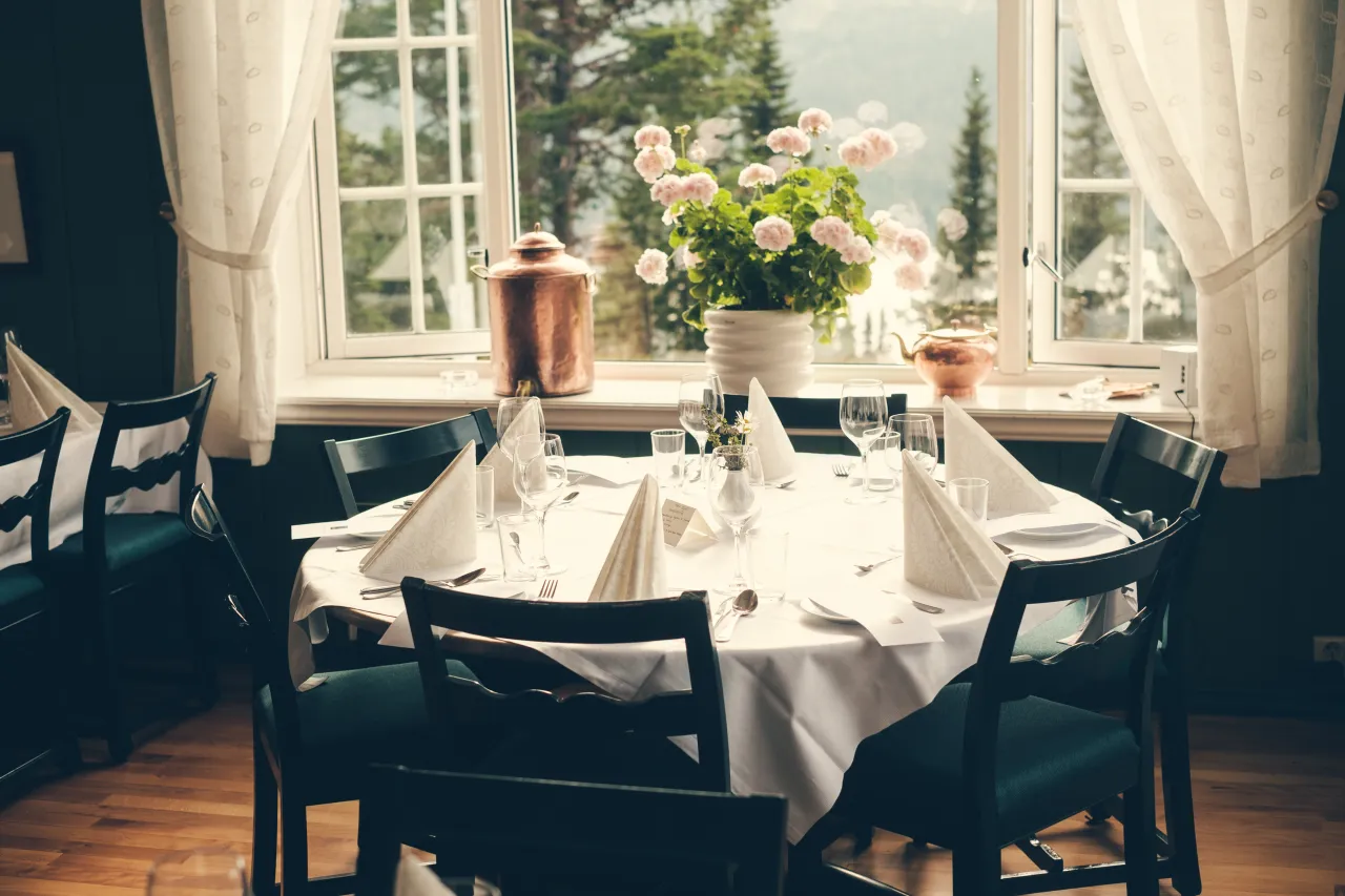 Restauranten er dekket til selskap, hvite duker, runde hovedbord med kobber og blomstrende pelargonia i vinduskarmen